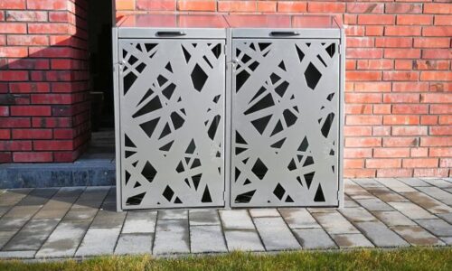 Zabudowa kosza 120l z geometrycznym wzorem na tle czerwonej ściany klinkierowej i zielonej trawy.